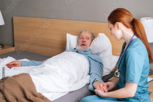 Smiling female nurse checking pulse on wrist to sick senior male patient lying on bed in hospital room. Doctor checking mature patient pulse at home. Elderly man getting medical consultation.