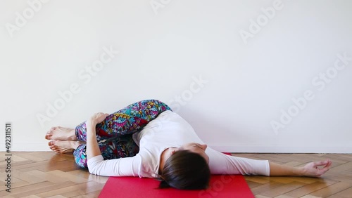 Caucasian young woman on Supine Spinal Twist yoga posture in a indoor room over red yoga mat and wood floor with white indoor room wall	
