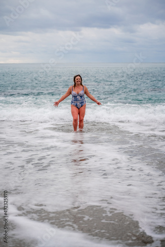 A plump woman in a bathing suit enters the water during the surf. Alone on the beach, Gray sky in the clouds, swimming in winter.