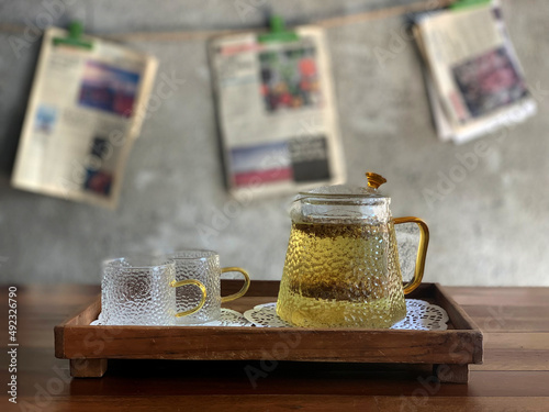 Herbal tea inside transparent cup  and luxury jar on wooden table. The decoration of newspaper hanging on hemp rope on cement wall. Tranquil space inside coffee cafe