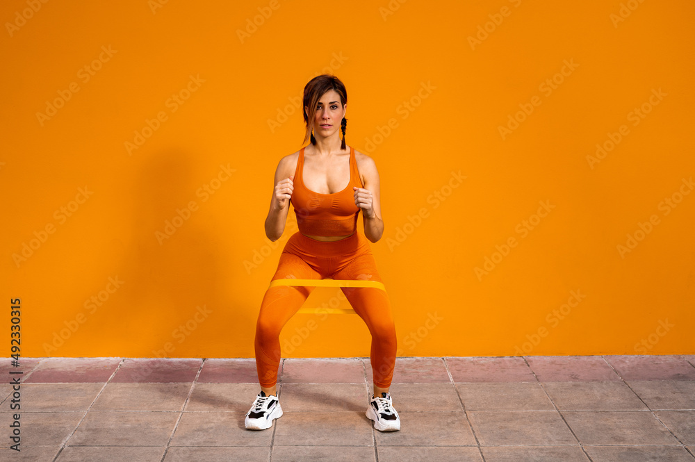 Atractiva mujer deportista haciendo ejercicios físicos con una banda elástica sobre un fondo de color naranja