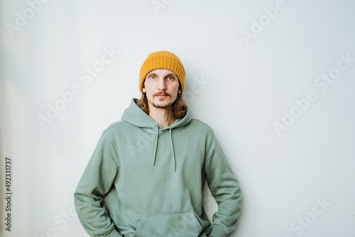 A portrait of a young man looks into the camera, a hairy guy in a yellow hat on his head stands against a white wall.