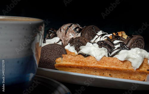Plate of belgian waffles with ice cream, cookies and chocolate sauce on dark background