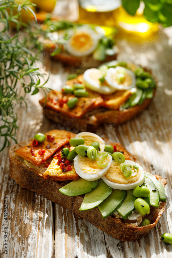 Vegetarian sourdough bread open face sandwiches with avocado, hard-boiled egg, tofu and edamame seasoned with herbs on a wooden background, close up view