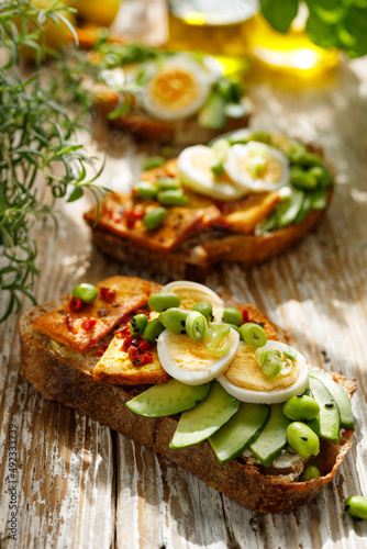 Vegetarian sourdough bread open face sandwiches with avocado, hard-boiled egg, tofu and edamame seasoned with herbs on a wooden background, close up view