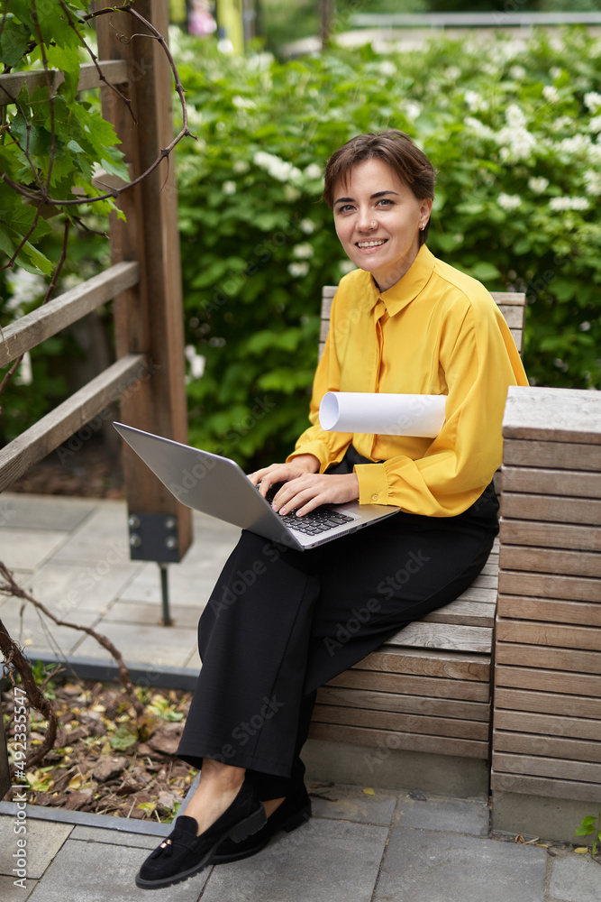 Fototapeta premium Business, finance industry or real estate concept. Smiling successful business lady in yellow blouse sitting on a bench outdoor in park using a laptop working looking at camera. High quality image