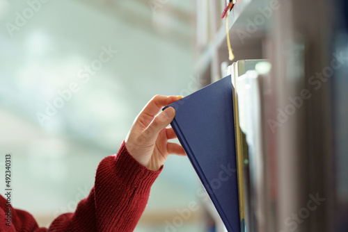 Female hand pulling book from bookshelf in public library in university, college or high school. Woman student take novel from bookcase in bookshop store, soft focus. Education and literature concept photo