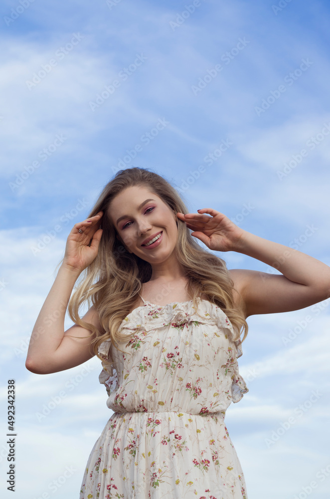 cheerful smiling blonde caucasian young woman in a summer dress with floral print looking at the camera on the background of the blue sky with clouds