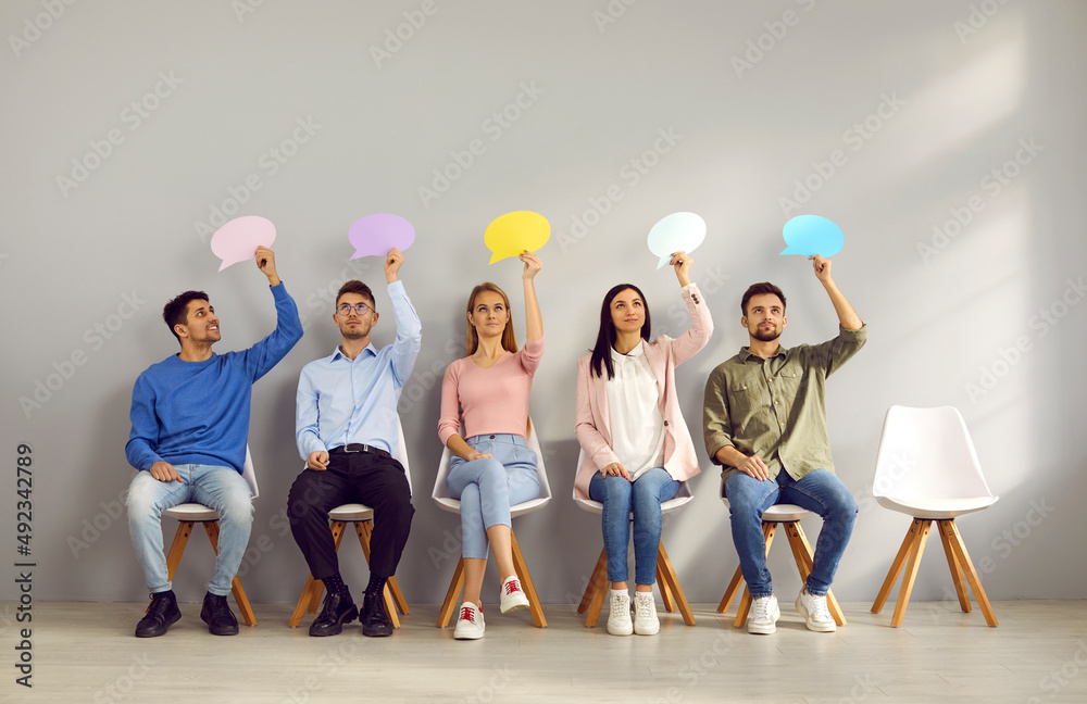 © Studio Romantic - Group of people sitting on row of chairs, thinking about something and holding empty colorful mock up speech bubbles. Young male and female company employees taking part in survey and sharing opinions