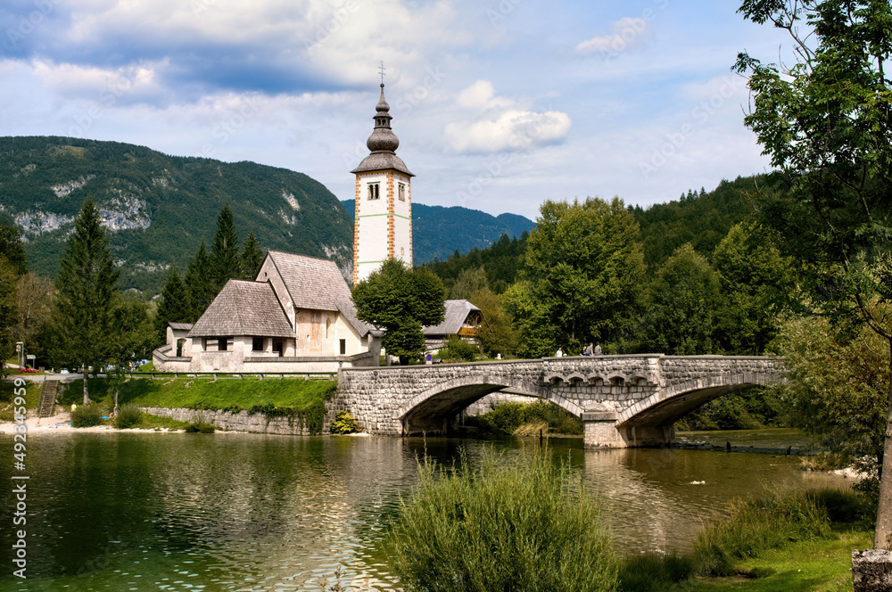Church with a tall tower and an old stone bridge over the water at Lake Bohinj in Slovenia.
