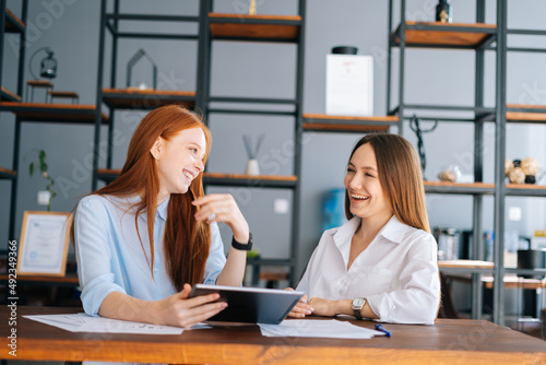 Front view of two laughing young businesswomen working using digital tablet at meeting desk with job documents at office. Business female colleagues using touchscreen computer for project discussion