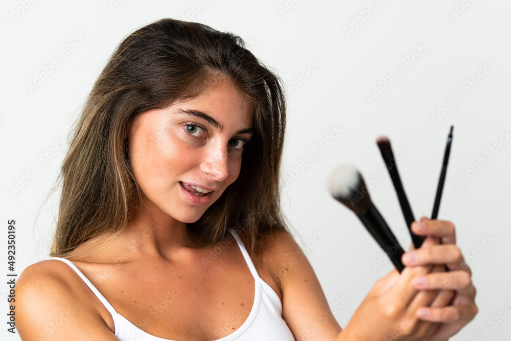 Young caucasian woman isolated on white background holding makeup brush and whit happy expression Young caucasian woman isolated on white background holding makeup brush and whit happy expression