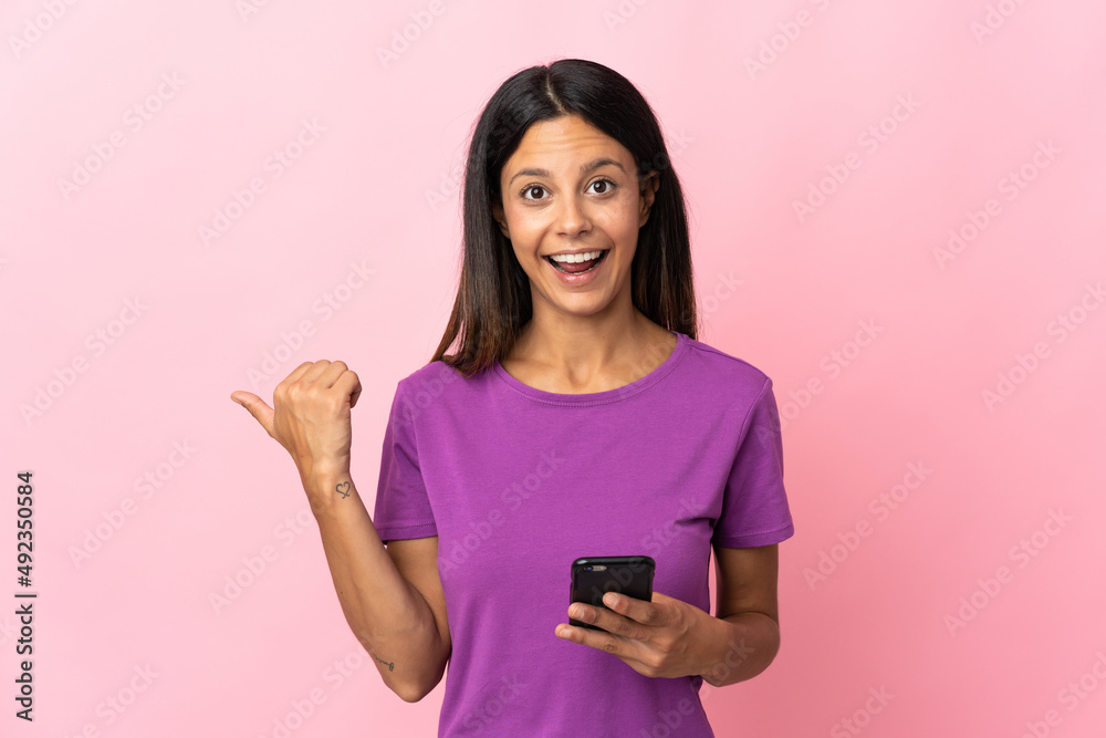 Caucasian girl isolated on pink background using mobile phone and pointing to the lateral Caucasian girl isolated on pink background using mobile phone and pointing to the lateral