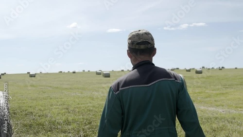 Male farmer man in cap and green workwear walking through green field near haystacks. Agriculture healthy food business farming concept. Rear view, Stedycam shot photo