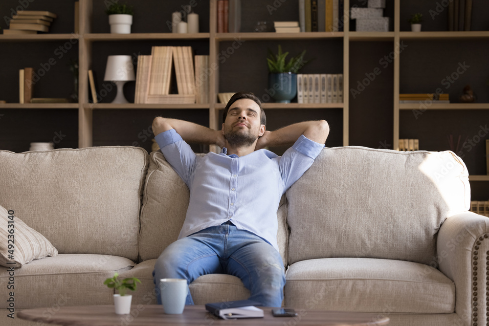 Peaceful millennial guy relax at quiet cozy living room dream enjoy tranquility peace of mind. Calm mindful young man take break from work study recline on sofa in comfortable pose keep eyes closed