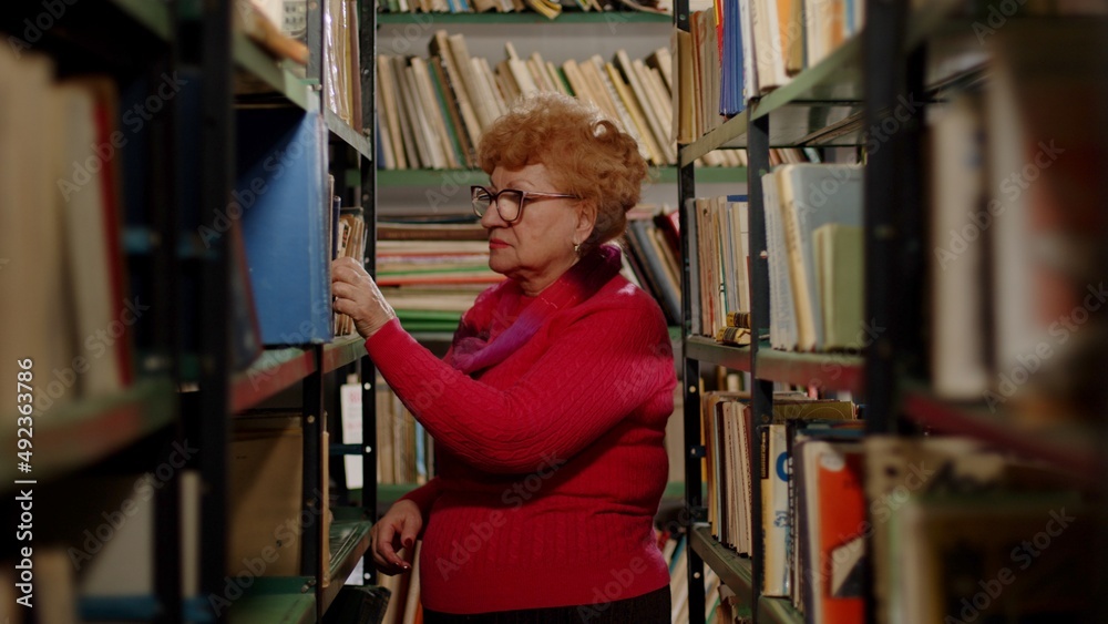 An elderly friendly woman in glasses is looking for a book on a bookshelf. Self-education An elderly friendly woman in glasses is looking for a book on a bookshelf. Self-education