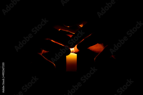 Hands are illuminated from the flame of a candle on a black background. The concept is the preservation of peace, warmth. ritual in magic. memory ritual.