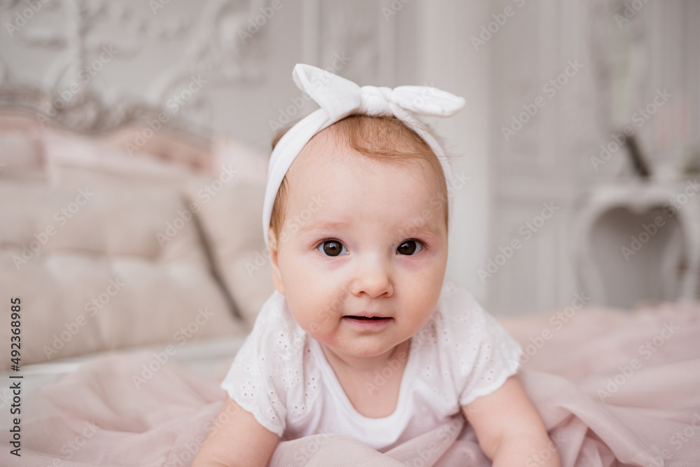 portrait of a Caucasian baby girl crawling on a bed in a room. Brown-eyed little girl in a white headband portrait of a Caucasian baby girl crawling on a bed in a room. Brown-eyed little girl in a white headband