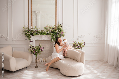 A Caucasian brunette mom is sitting on the couch in a beautiful room with a baby girl. Mom playing with her baby daughter