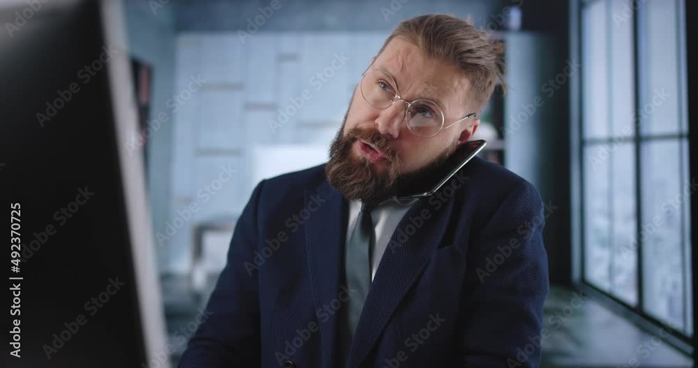 Serious bearded man in formal clothes typing on computer and talking on mobile during working process at office. Concept of people, business and technology. Serious bearded man in formal clothes typing on computer and talking on mobile during working process at office. Concept of people, business and technology.