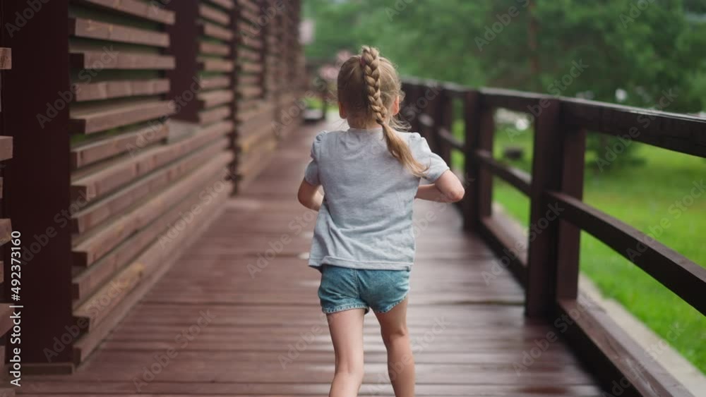Little girl with blonde plait runs playing along empty veranda deck at suburban cottage house on spring day backside view slow motion Little girl with blonde plait runs playing along empty veranda deck at suburban cottage house on spring day backside view slow motion
