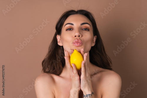 Beauty portrait of topless woman with perfect skin and natural make-up on a beige background holds citrus lemon, vitamins c for skin