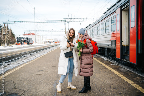 two girls on the station platform with flowers happily looking into a smartphone photo
