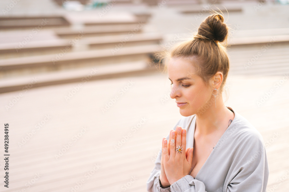 Close-up face of meditative young woman practicing yoga performing namaste pose with closed eyes outside in city park. Beautiful serene female meditating and doing yoga posture outdoors alone. Close-up face of meditative young woman practicing yoga performing namaste pose with closed eyes outside in city park. Beautiful serene female meditating and doing yoga posture outdoors alone.