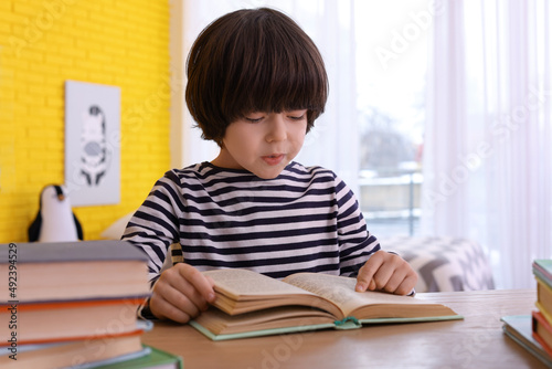 Cute little boy reading book at table in room
