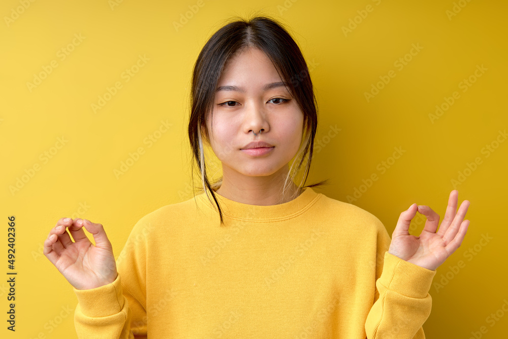 calm confident asian or chinese woman makes zen nirvana gesture meditates looking at camera practices yoga, brunette female wears casual yellow jumper, posing isolated over yellow background. calm confident asian or chinese woman makes zen nirvana gesture meditates looking at camera practices yoga, brunette female wears casual yellow jumper, posing isolated over yellow background.