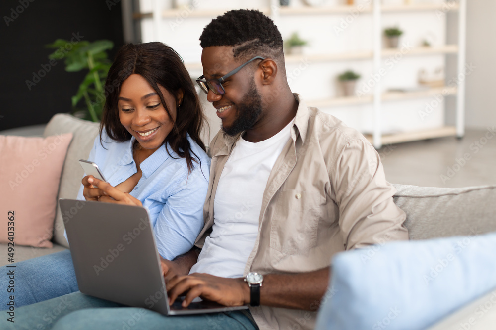 Happy black couple sitting on couch, using cellphone and pc