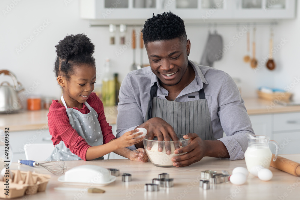 Little black girl adding eggs while her daddy making dough Little black girl adding eggs while her daddy making dough