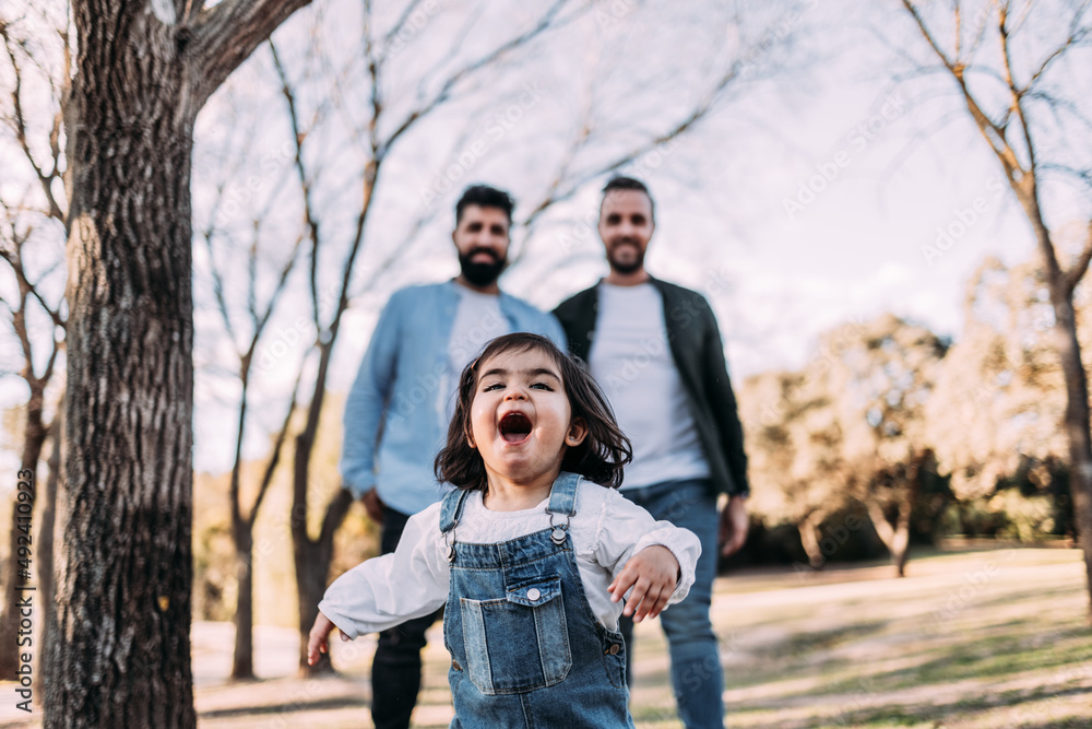 A little girl running happily through the park followed by her parents. Gay family concept. A little girl running happily through the park followed by her parents. Gay family concept.