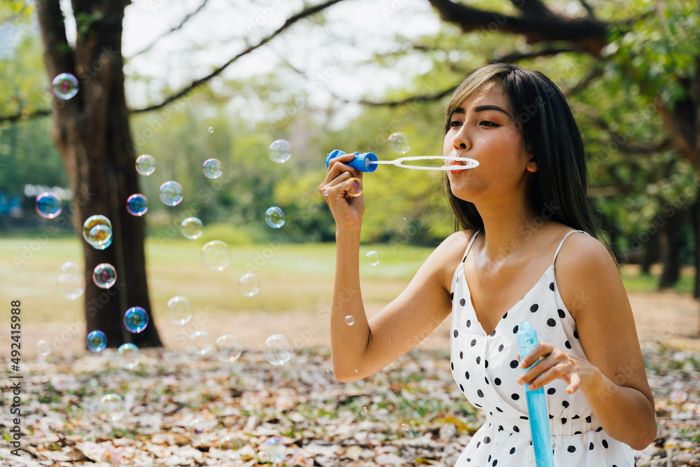 South East Asian ethnic 20s woman blowing air bubbles in the park Young Happy girl wears stylish dress with trees in background. Outdoor summer weekend activity - with copy space South East Asian ethnic 20s woman blowing air bubbles in the park Young Happy girl wears stylish dress with trees in background. Outdoor summer weekend activity - with copy space