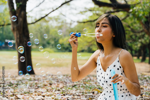 South East Asian ethnic 20s woman blowing air bubbles in the park Young Happy girl wears stylish dress with trees in background. Outdoor summer weekend activity - with copy space