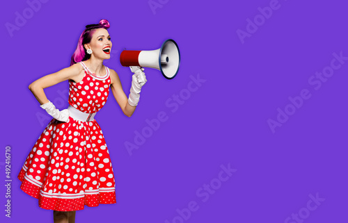 Beauty red haired woman holding megaphone, shout something. Girl in pin-up style dress in polka dot, isolated over purple violet background. Caucasian model posing in retro fashion vintage concept.