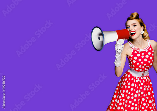 Beauty blond haired woman holding megaphone, shout something. Girl in red pin up style dress in polka dot, isolated over purple violet background. Retro and vintage studio concept.