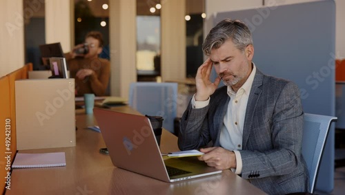 Portrait of aucasian businessman working on a laptop in the office having a terrible headache during working hours. Overworker and healthcare concept. photo