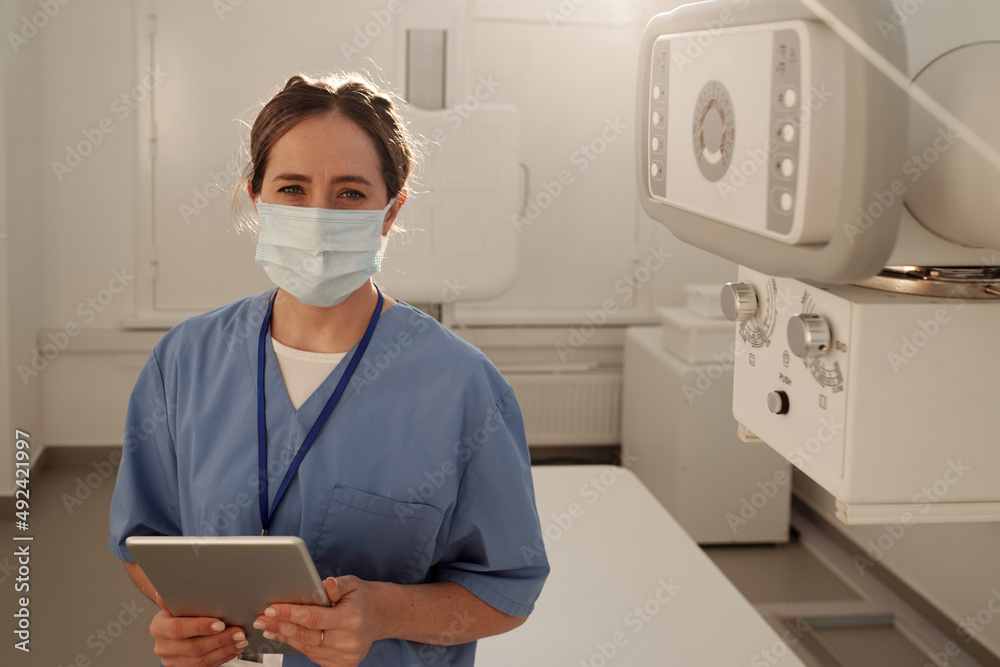 Young contemporary female radiologist in protective mask holding digital tablet while consulting clients online in hospital