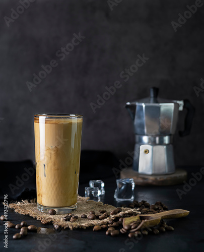 Transparent glass with cold coffee on a canvas napkin. Coffee pot on a wooden stand in the background. Ice cubes and coffee beans on a black table