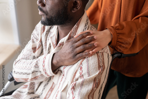 Young African American man in wheelchair covering hand of female caregiver on his shoulder while looking through window