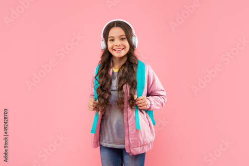 happy child listen music in headphones and carry backpack on pink background, school