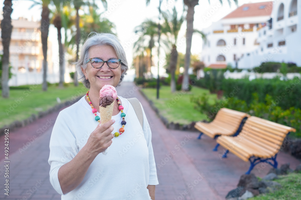 Fototapeta premium Senior woman smiling eating ice cream of chocolate and strawberry looking at camera. Happy female retiree enjoying sweet food in public park
