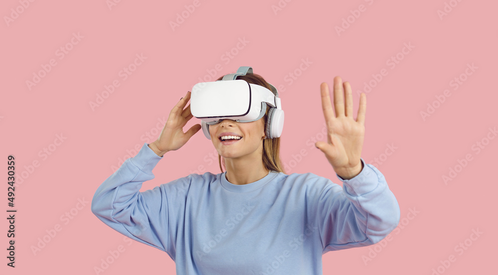 Happy young woman in VR headset experiencing virtual reality. Teen female gamer student in VR glasses learning about world while exploring new exciting videogame. Studio shot on pastel pink background