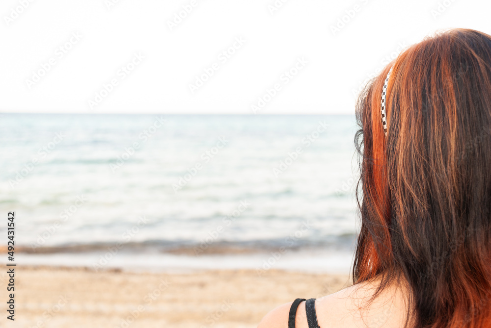 Young woman with red and black long hair standing at the beach, looking at the sea, rear view, horizontal Young woman with red and black long hair standing at the beach, looking at the sea, rear view, horizontal