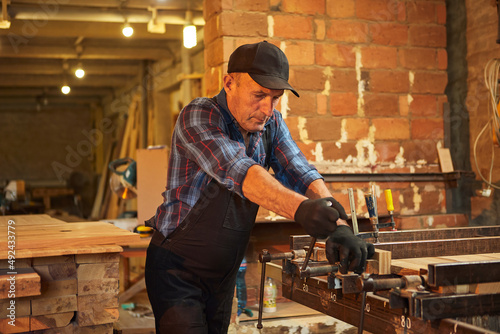 Portrait of a senior carpenter in uniform gluing wooden bars with hand pressures at the carpentry manufacturing