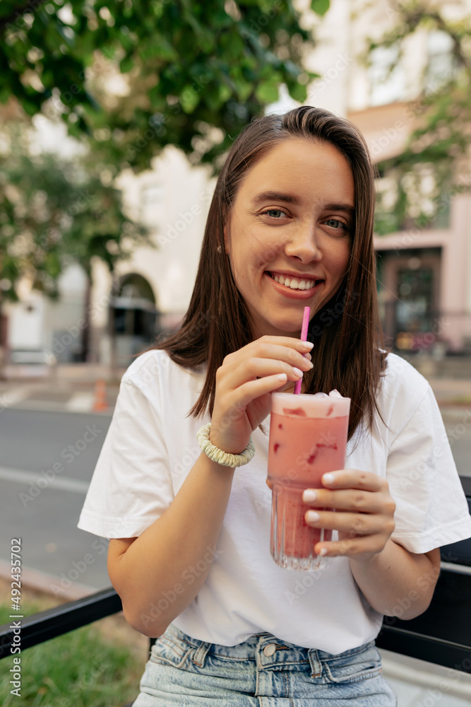 Charming lovely smiling woman with dark hair is drinking summer smoothie and smiling at camera while resting in open air cafe on blur city background Charming lovely smiling woman with dark hair is drinking summer smoothie and smiling at camera while resting in open air cafe on blur city background