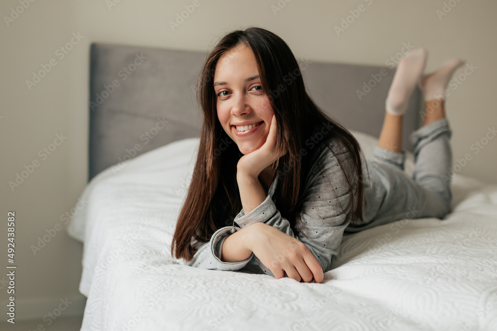 Early morning in modern apartment of cheerful attractive girl sleeping on white bed. Charming young woman is resting, smiling and looking at camera Early morning in modern apartment of cheerful attractive girl sleeping on white bed. Charming young woman is resting, smiling and looking at camera
