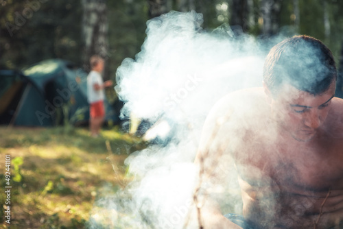 Man in smoke during cooking on fire outdoors on field kitchen in camping site as camping lifestyle