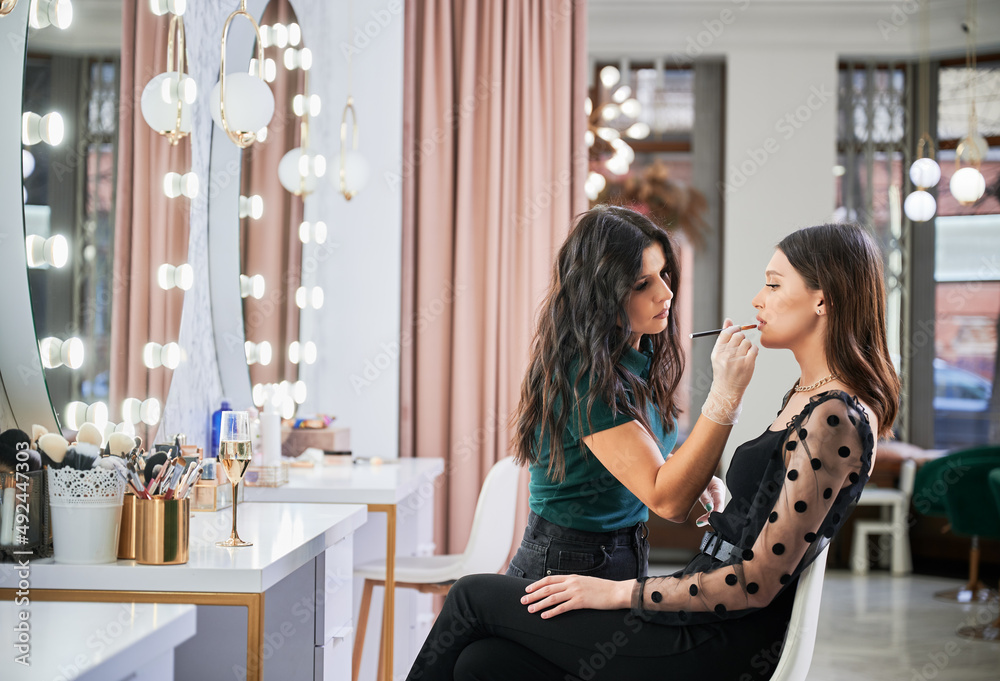 Makeup artist doing professional in visage studio. Stylish woman sitting at dressing table while female beauty specialist in gloves applying lipstick on client lips with cosmetic brush. Photos | Adobe